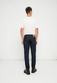 Jeans din denim albastru închis cu o croială dreaptă, având două buzunare la spate și o etichetă din piele bej pe talie. Tricou alb simplu.