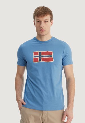 Jeune homme portant un t-shirt bleu avec un motif du drapeau norvégien rouge, blanc et bleu, debout, une main dans la poche d'un pantalon beige.