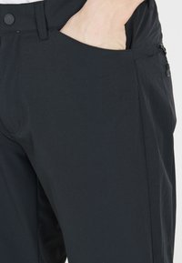 Mustat housut, jotka on valmistettu joustavasta kankaasta, sisältävät litteän etuosan, sivutaskun ja vetoketjullisen taskun turvallista säilytystä varten.