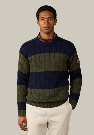 Mann trägt einen marineblau und olivgrün gestreiften Zopfpullover über einem karierten Hemd mit beiger Hose vor beigem Hintergrund.