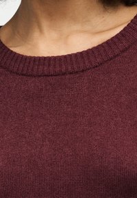 Gros plan d'une personne portant un pull à côtes avec un col rond en tissu de laine bordeaux foncé.