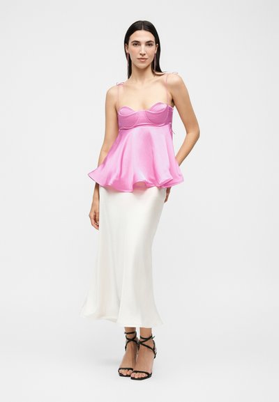 Γυναίκα με λαμπερό ροζ αμάνικο τοπ με λεπτές τιράντες και λευκή μέση φούστα, μαύρα ψηλοτάκουνα πέδιλα με λουράκια, ποζάρει με το ένα χέρι στη μέση.