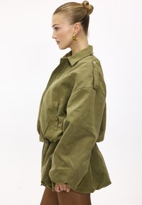 Femme aux cheveux blonds attachés en chignon, portant une veste oversize vert olive et des boucles d'oreilles créoles en or, debout de profil devant un fond blanc.