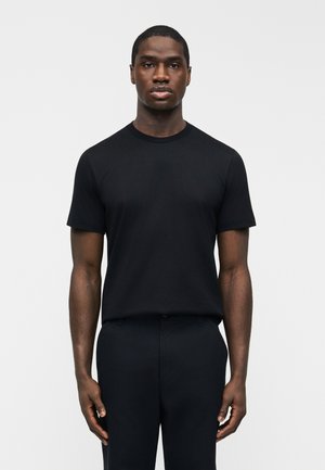 Άνδρας που στέκεται απέναντι, φορώντας εφαρμοστό μαύρο κοντομάνικο μπλουζάκι και μαύρο παντελόνι μπροστά σε απλό ανοιχτόχρωμο φόντο.