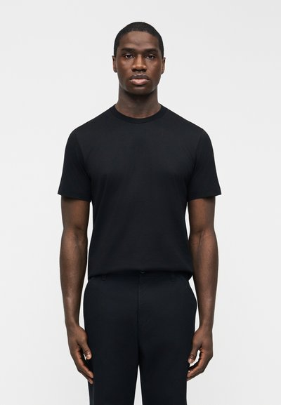 Homme debout, faisant face à l'avant, portant un t-shirt noir ajusté à manches courtes et un pantalon noir, sur un fond clair uni.
