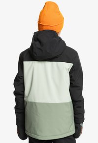 Färgblockerad jacka med svarta, ljusgröna och ljusgrå sektioner; justerbar huva; texturerat material; bärs med en orange mössa.