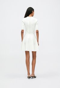 Vestido blanco con parte superior ajustada y falda en forma de A, mangas cortas, textura acanalada y detalle de cremallera en la parte posterior. Llevado con tacones negros.