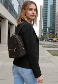 Zaino in nylon nero con forma arrotondata, cerniere dorate e una tasca frontale. Indossato su una spalla, abbinato a una giacca nera e jeans blu.