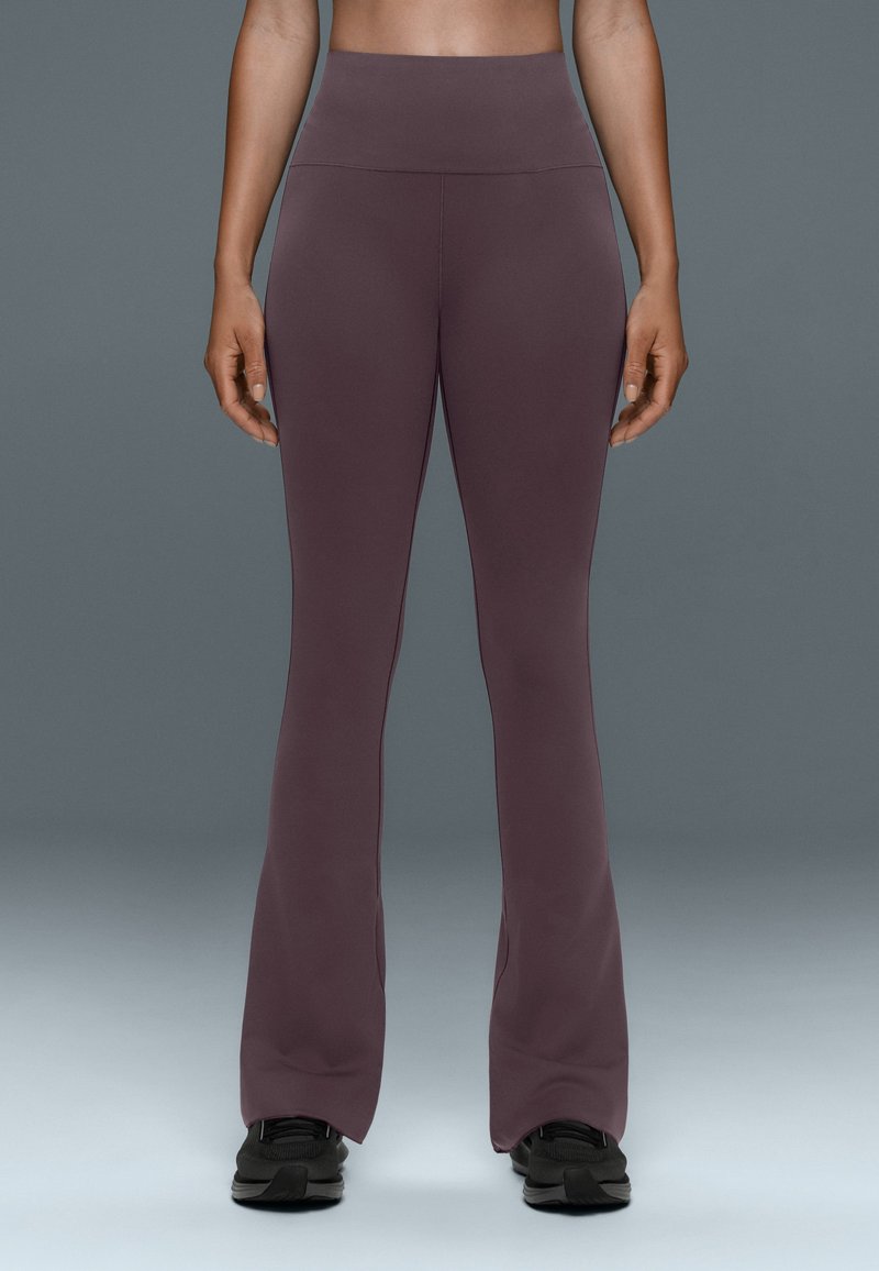 Personne portant des leggings violets taille haute évasés et des chaussures de sport noires, debout devant un fond gris uni.