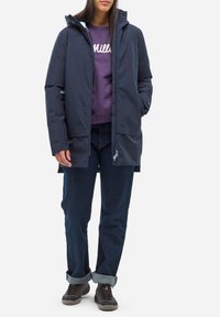 Manteau bleu marine résistant à l'eau avec capuche, fermeture éclair à l'avant et poches latérales, superposé sur un sweat-shirt violet et un jean en denim foncé.