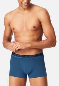 Smiling man wearing blue patterned boxer briefs with "Uncover" waistband, standing against a plain light background.
