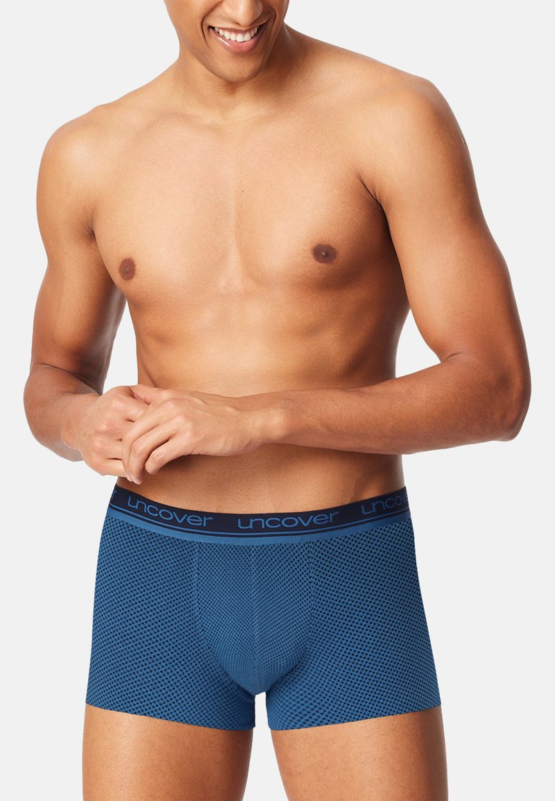Smiling man wearing blue patterned boxer briefs with "Uncover" waistband, standing against a plain light background.