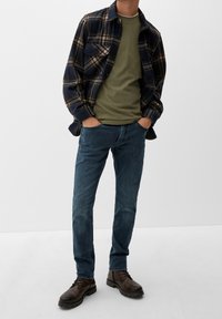 Camicia di flanella a quadri nei colori blu navy e beige, t-shirt girocollo verde oliva, jeans blu e stivali in pelle marrone con suola testurizzata.