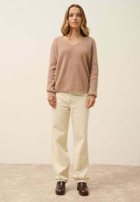 Jasno beżowy sweter w serek z miękkiego dzianiny, zestawiony z kremowymi szerokimi spodniami. Modelka ma na sobie ciemnobrązowe botki na obcasie, stojąc na neutralnym tle.