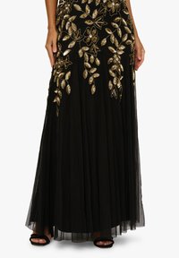 Černá sukně až na zem s zlatou květinovou výšivkou a plisovanou tylem nahoře, nošena s černými sandály na otevřenou špičku.