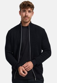 Musta teksturoitu vetoketjullinen takki, jossa korkeahko kaulus. Takissa on kontrastivärinen vetoketju ja resoriset hihansuut. Päällä musta t-paita.