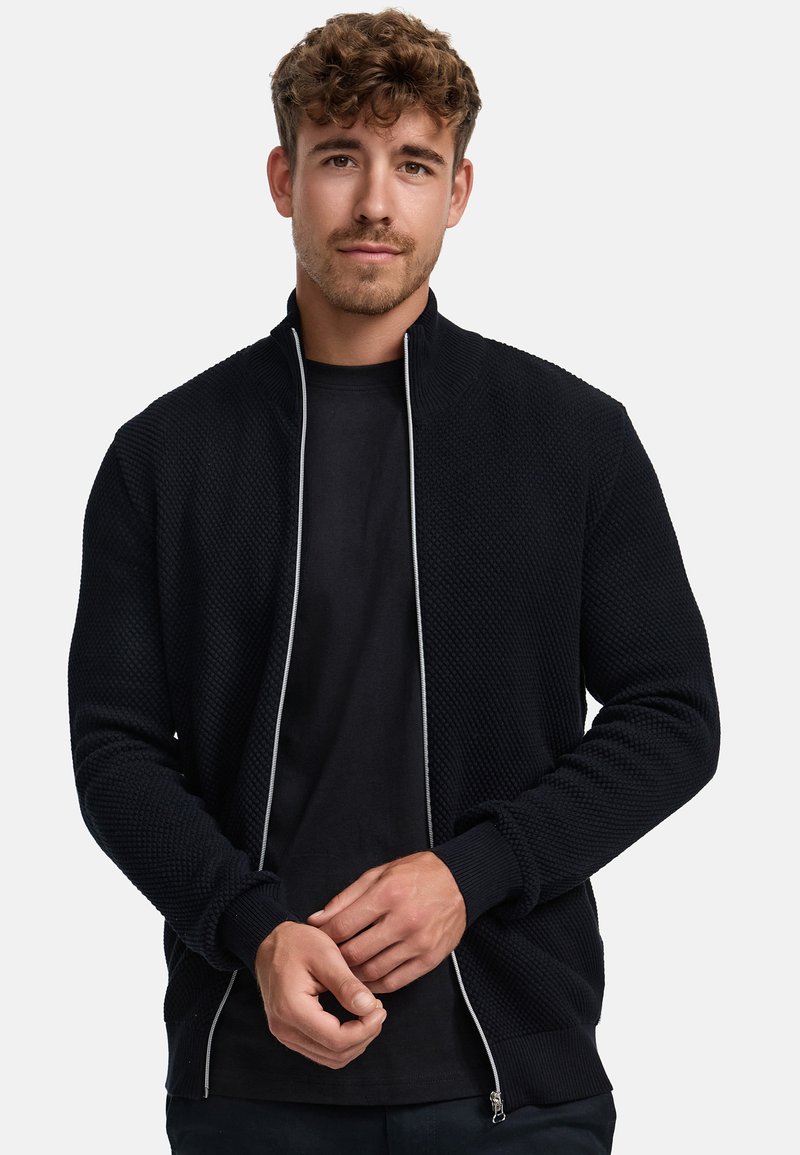 Musta teksturoitu vetoketjullinen takki, jossa korkeahko kaulus. Takissa on kontrastivärinen vetoketju ja resoriset hihansuut. Päällä musta t-paita.