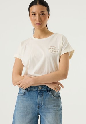 Off-white cotton t-shirt with rolled short sleeves, featuring embroidered text accents in beige: “Amore In Fiore” and “Love Garcia.” Blue jeans.