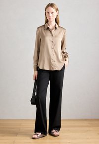 Kvinna står och håller en svart väska, klädd i en beige satinknappad skjorta, svarta vida byxor och metalliska sandaler på ett trägolv.