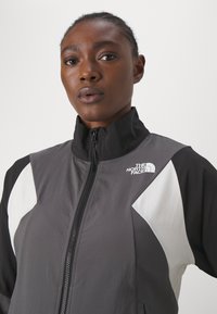 Giacca zip-up grigia e nera con accenti bianchi, colletto alto e logo sul lato sinistro. Materiale leggero con una texture liscia.