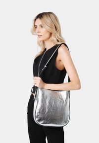 Borsa a spalla metallicamente argento con una texture liscia, forma curvata e rifiniture nere. Cinghia regolabile e dettagli con cerniera sulla parte frontale.