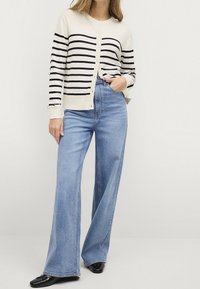 Cardigan rayé en crème et marine, associé à un jean bleu à taille haute et jambes larges. Des chaussures plates noires complètent l'ensemble.