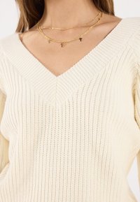 Pull en maille côtelée crème avec un col en V profond. Colliers en chaînes dorées superposés avec de petits pendentifs croix. Texture subtile et design léger.