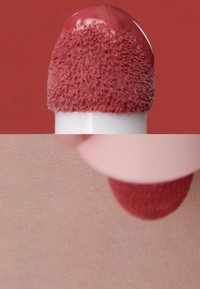 Aplicador de maquillaje líquido rosado con una punta de espuma texturizada, aplicado sobre piel clara. El fondo es de color rosa apagado sólido.