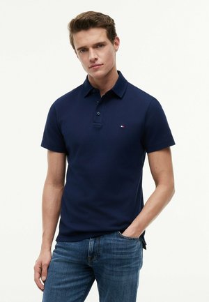 Ung mand iført navyblå poloshirt og blå jeans, stående med den ene hånd i lommen mod en ensfarvet lys baggrund.