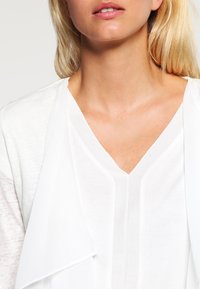White layered cardigan with a soft texture. Features a V-neckline and draped front. Smooth fabric contrasts with slightly textured sleeves.