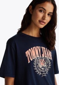 Mørkeblå bomulls T-skjorte med "TOMMY JEANS" grafisk logo i ferskenfarge, med et tiger- og laurbærdesign trykt på midten. Korte ermer.