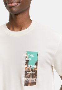 Cremefarbenes Baumwoll-T-Shirt mit einem aufgedruckten rechteckigen Grafikmotiv in Türkis und fotografischen Elementen, mit dem Text "ON THE ROAD TO CALIFORNIA."