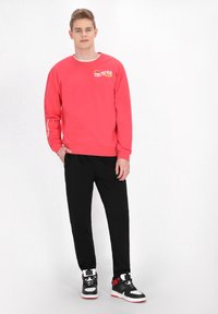 Felpa rossa con accenti grafici bianchi e gialli, abbinata a pantaloni jogger neri. Sneakers in bianco e nero con dettagli rossi completano il look.