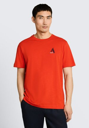 Mężczyzna ubrany w jaskrawopomarańczowy t-shirt z krótkim rękawem i małym granatowym logo żaglówki na piersi, w ciemnych spodniach, stojący na jednolitym tle.