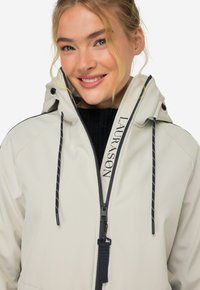Giacca beige chiaro realizzata in materiale morbido, con cappuccio, chiusura a zip, cordini neri e dettagli scuri a contrasto.