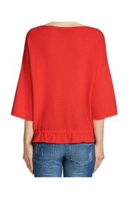 Rotes, locker sitzendes Oberteil mit einem strukturierten Waffelstrickdesign und weiten Ärmeln, das einen gerafften Saum hat. Kombiniert mit hellblauen, verwaschenen Jeans.
