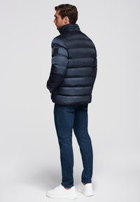 Mørkeblå pufjakke med høj krave, der har horisontale quiltede sektioner; båret med mørke jeans og hvide sneakers, set fra ryggen.