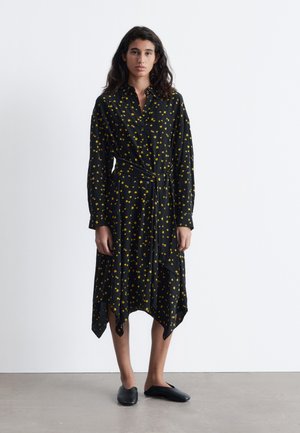 Schwarzes Hemd-Kleid mit gelbem Blumenmuster, Knopfverschluss, langen Ärmeln und asymmetrischem Saum mit spitzen Kanten. Getragen mit schwarzen Ballerinas.