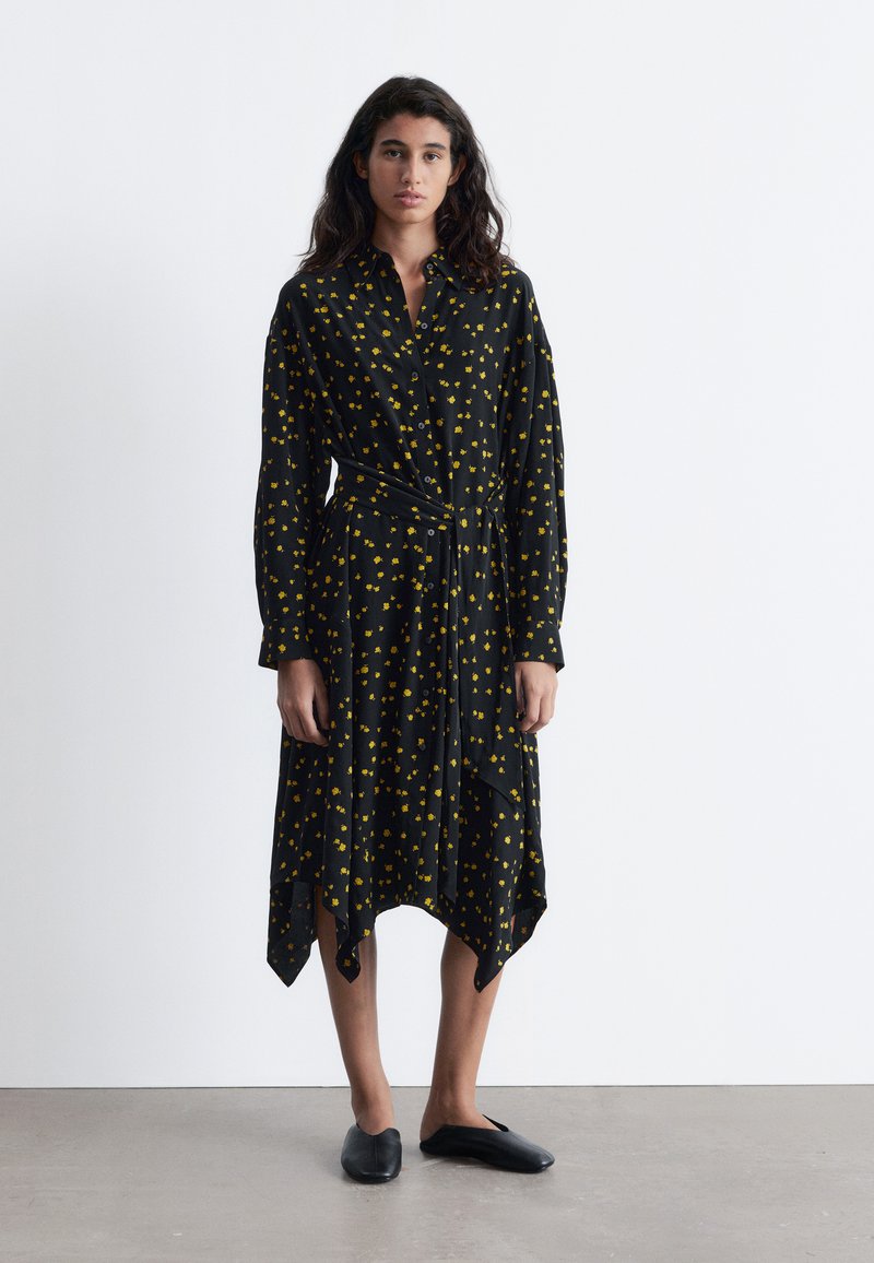 Schwarzes Hemd-Kleid mit gelbem Blumenmuster, Knopfverschluss, langen Ärmeln und asymmetrischem Saum mit spitzen Kanten. Getragen mit schwarzen Ballerinas.