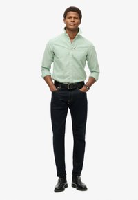 Camisa de botones verde claro con bolsillo en el pecho, vaqueros azul oscuro y botas de cuero negras. El modelo está de pie con las manos en los bolsillos frente a un fondo blanco.