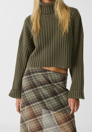 Un pull en tricot côtelé vert olive avec un col montant et des manches évasées, associé à une jupe en tulle à carreaux aux tons marron et beige.