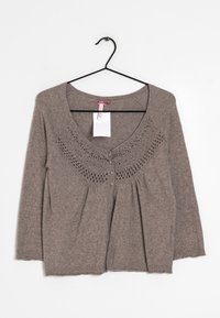 Brun stickad cardigan med långa ärmar, dekorativt virkade mönster runt halsringningen och knappar framtill, hängande på en svart galge.