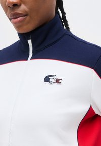 Ženska, oblečena v belo, mornarsko modro in rdečo jakno z zadrgo, z logotipom krokodila na prsih in pletenicami, ki so vidne za ramom.