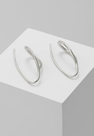 Paire de boucles d'oreilles en fil d'argent torsadé reposant sur un cube blanc sur un fond gris clair.