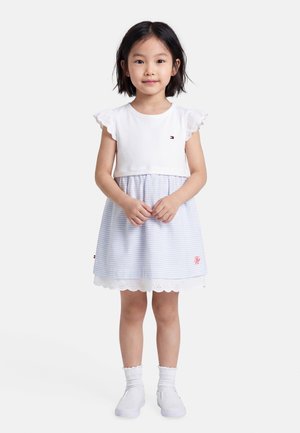 Mladá dívka stojí, má na sobě bílé šaty s volánkovými rukávy a světle modrými pruhy, bílé ponožky a bílé nazouvací boty na jednobarevném pozadí.