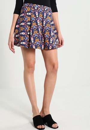 Vrouw draagt een geplooide rok met geometrisch patroon in oranje, roze, wit en donkerblauw, gecombineerd met zwarte open-toe muilhakken.