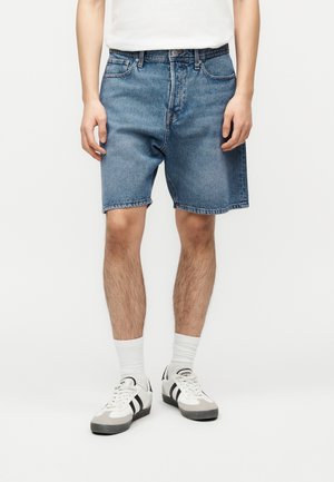 Person trägt blaue Jeans-Shorts mittlerer Länge, weiße Sportsocken, weiße und schwarze Sneakers und ein weißes T-Shirt und steht vor einem schlichten Hintergrund.
