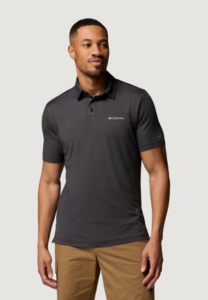 Homme portant un polo à manches courtes Columbia gris foncé et un pantalon beige, debout devant un fond clair uni.