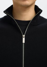 Gros plan d'une personne portant un pull en maille noir zippé sur un t-shirt noir avec la fermeture éclair partiellement fermée.