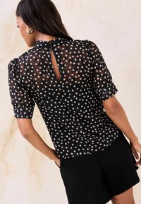 Mustad täpiline pluus läbipaistvast kangast, lühikeste muhklikute varrukatega ja tagaküljel võtmeauguga detail. Sile tekstuur täiendab stiilset kujundust.